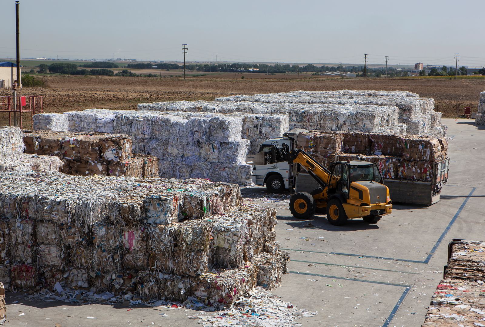 Recovery yard stacked with bales of recovered cardboard and mixed papers, a loader working between the rows